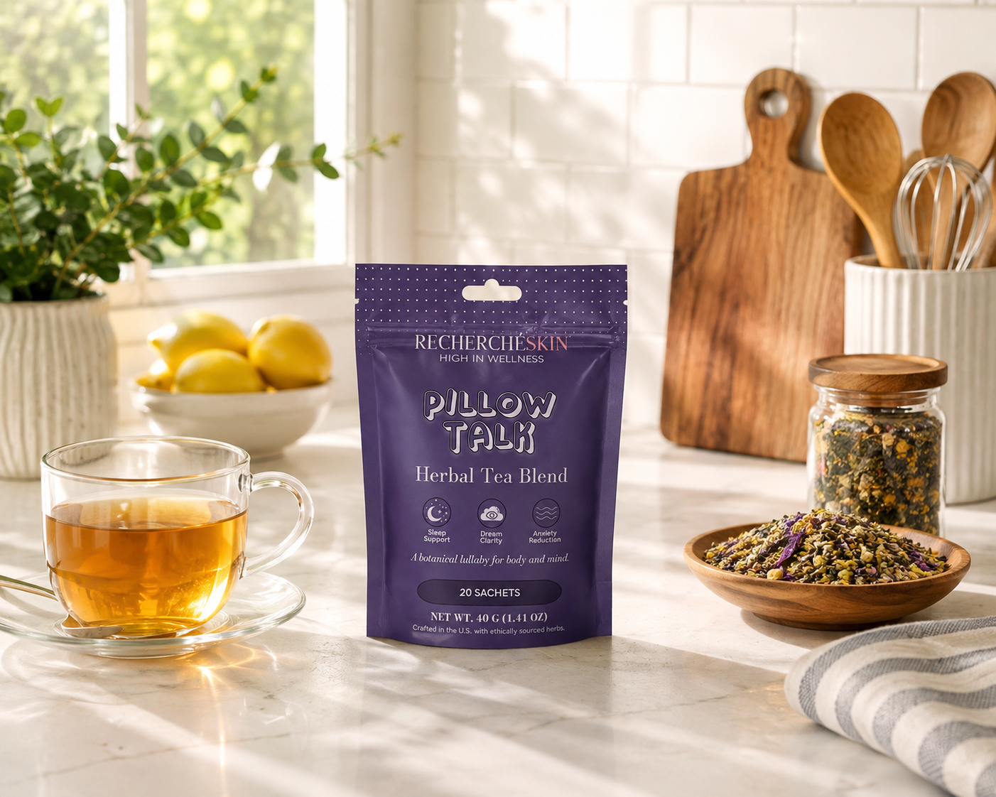 A purple stand-up pouch of Recherche Skin Pillow Talk Herbal Tea Blend sits on a bright kitchen counter, surrounded by a glass cup of amber tea, loose dried flowers in a bowl, and wooden kitchen tools in the background.